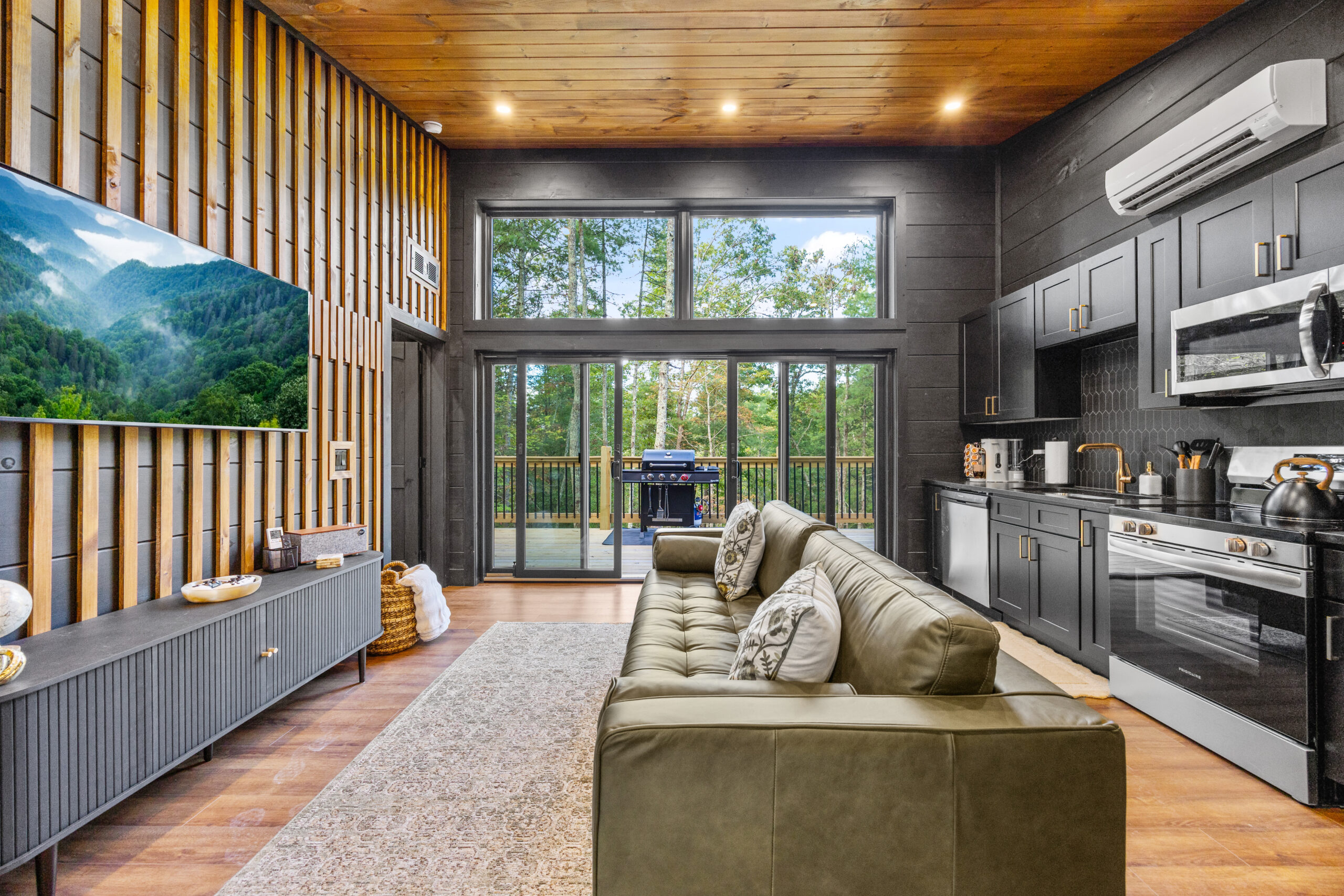 A high-end, open-concept living room and kitchen area in a luxury tiny cabin, featuring a flat-screen TV, modern furnishings, and a large glass door showcasing a panoramic view of the lush Smoky Mountains forest.