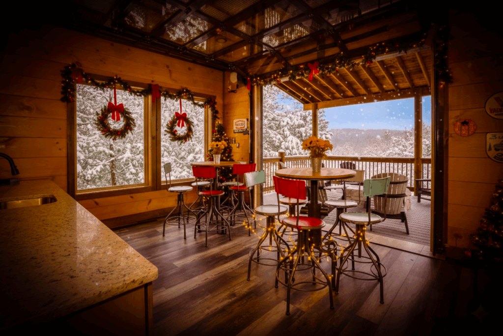 Image shows a beautiful, cozy, and comfortable indoor bar, with access to the balcony/terrace, features a garage-like roll door that connects the outside terrace with the inside bar. The bar is adorned with Christmas decorations, looks cozy, and it is snowy outside. Perfect place to spend the holidays.