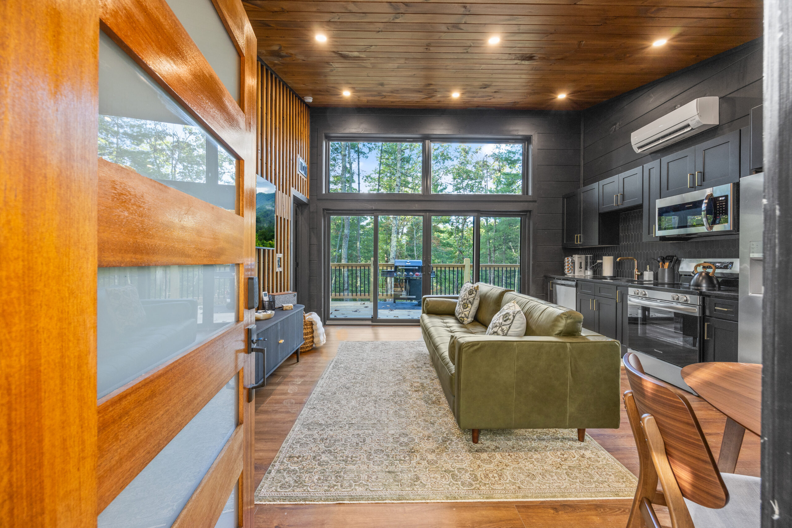 View of the living room space, kitchen, and terrace seen from the front door of a Contemporary Luxury Mountain Cabin. Showcases high-end design, appliances, custom-made decorations, interior design, and a glass terrace door. Built custom by the best builders in the Smoky Mountain, Pigeon Forge, Gatlinburg, Sevierville region.