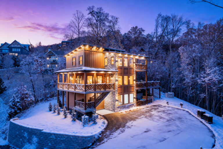 Exterior of a three-story luxury mountain cabin at dusk, glowing with warm interior lighting amidst a snowy landscape. A custom-engineered winter home by MCHB in the Smoky Mountains, Sevierville, TN.