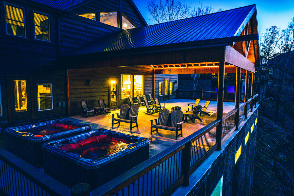 Rear view of a luxury mountain home by MCHB at dusk, featuring a massive wooden deck with two hot tubs and a glowing interior. A premium custom build in the Smoky Mountains, TN.