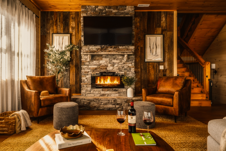 Cozy luxury living room in an MCHB custom cabin featuring a stone fireplace, leather seating, and wine glasses on a rustic center table. High-end interior design in the Smoky Mountains.