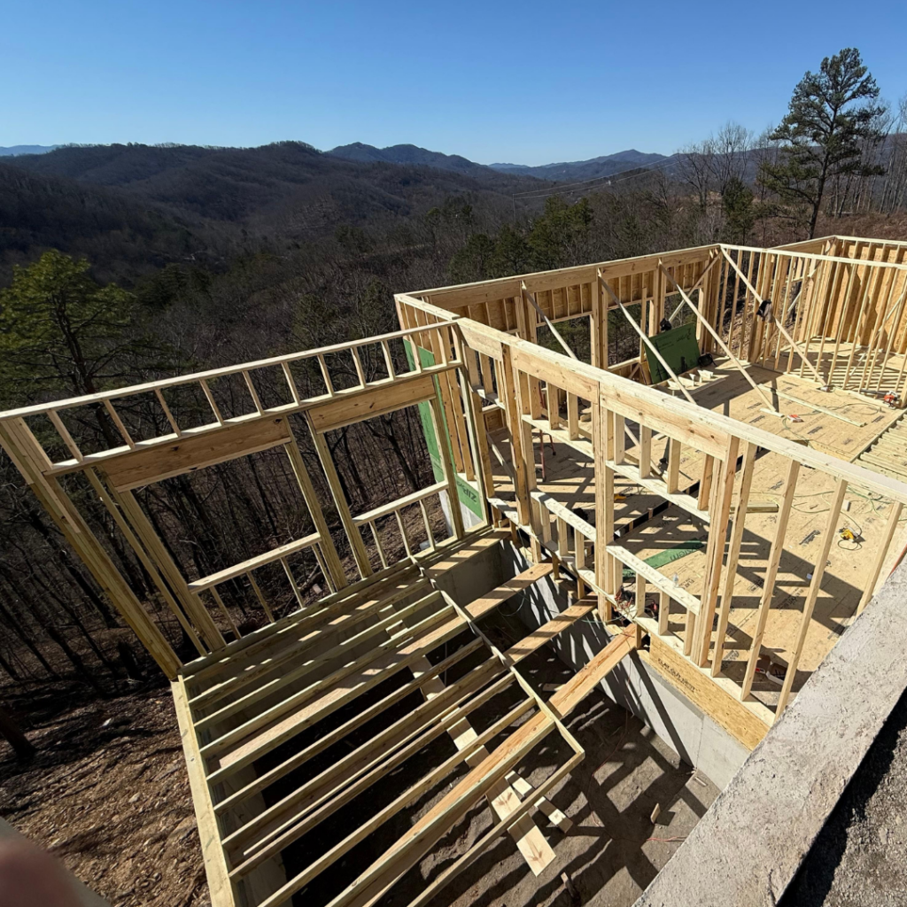 Active construction site of an MCHB luxury mountain home in Sevierville, TN. Features a bulldozer performing site grading and foundation prep under a clear blue sky in the Smoky Mountains.