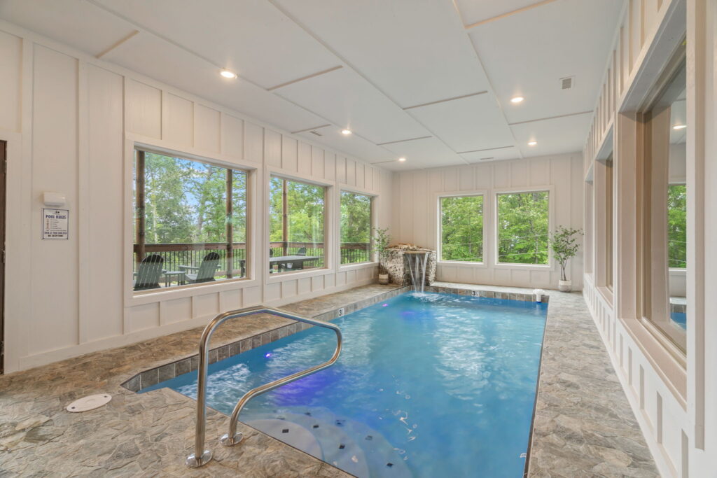 Luxury indoor heated pool inside Shell Mountain Sunrise cabin. Features modern stone tiling, wood accents, and large windows with Smoky Mountain views, engineered by MCHB.