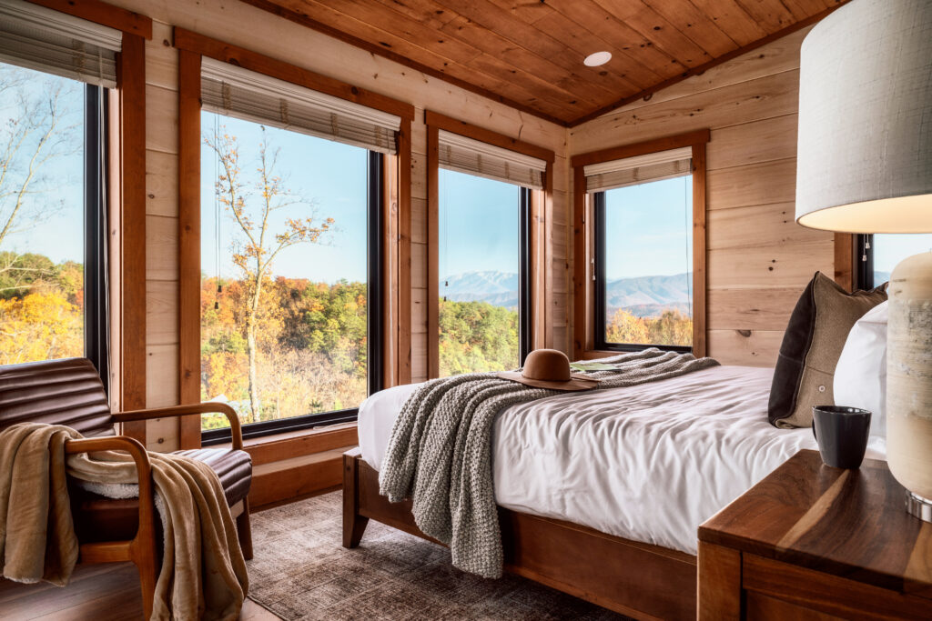 MCHB luxury modern cabin bedroom in Gatlinburg, TN. Features a custom-made bed, high-end linens, and two large, floor-to-ceiling windows framing a stunning panoramic view of the Smoky Mountains.