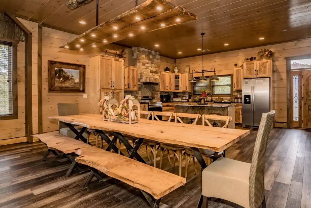 Integrated dining and kitchen area in a rustic MCHB mountain home. Features a large wooden dining table, marble countertops, crafted wood finishes, and modern artistic lighting in the Smoky Mountains.