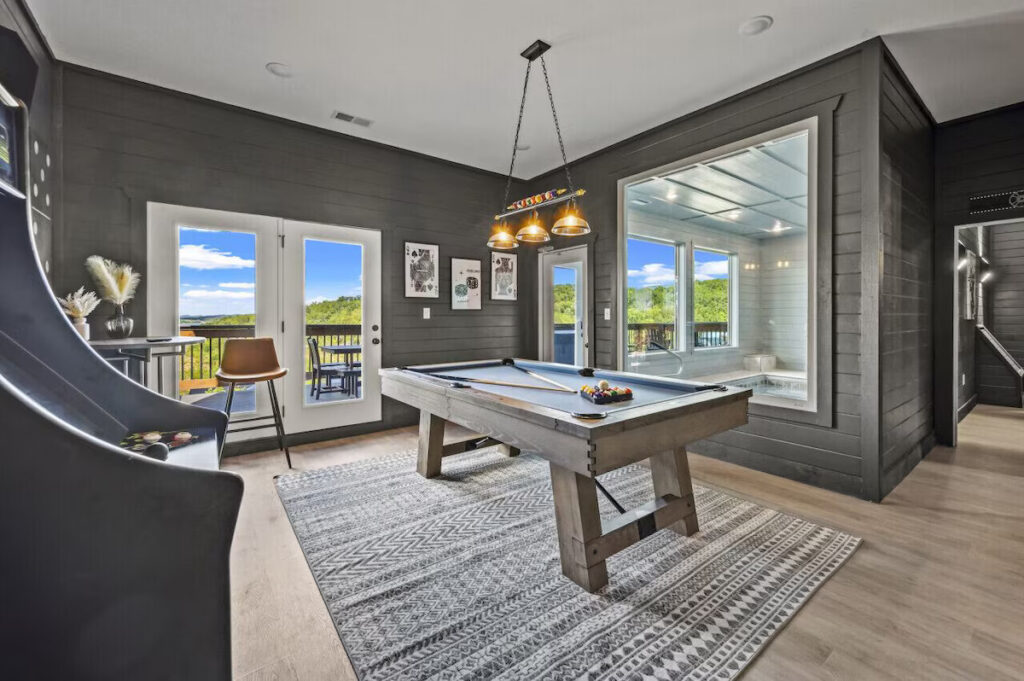 MCHB luxury mountain cabin game room with minimalistic aesthetics. Features a modern pool table, arcade games, and technology. Separated by a large glass window and door, it seamlessly integrates with an indoor swimming pool in Pigeon Forge, TN.