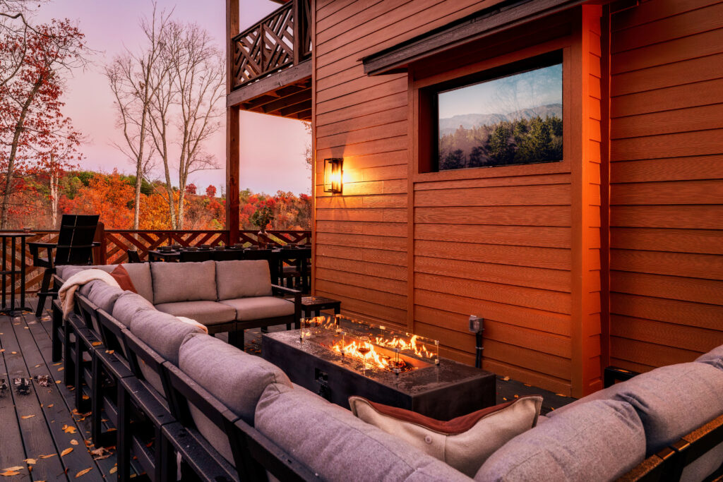 MCHB luxury mountain home balcony in Gatlinburg, TN. Features high-end outdoor furniture, an integrated fire pit, a stone outdoor entertainment center with TV, custom lighting, railings, and panoramic Smoky Mountain views in a modern-classic design.