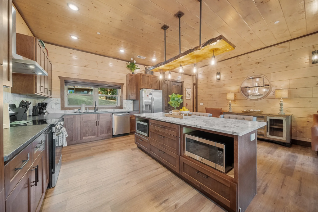 Wide-angle view of an MCHB modern-rustic kitchen in the Smoky Mountains. Features a multi-purpose marble counter, stove area, and back sink zone with artistic lighting and high-end appliances.