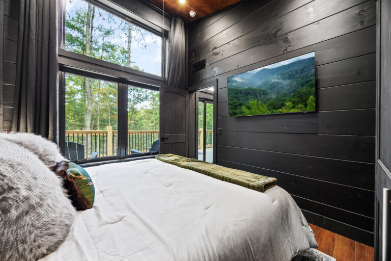 Master bedroom in a modern mini cabin featuring high-end linens, wall-mounted TV, and a glass door leading to a private balcony.