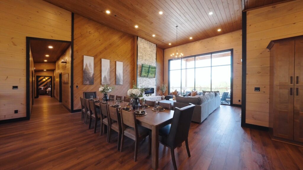 Side shot of an integrated open-concept living and dining room with vaulted ceilings and elegant lighting, leading to a floor-to-ceiling glass window wall.