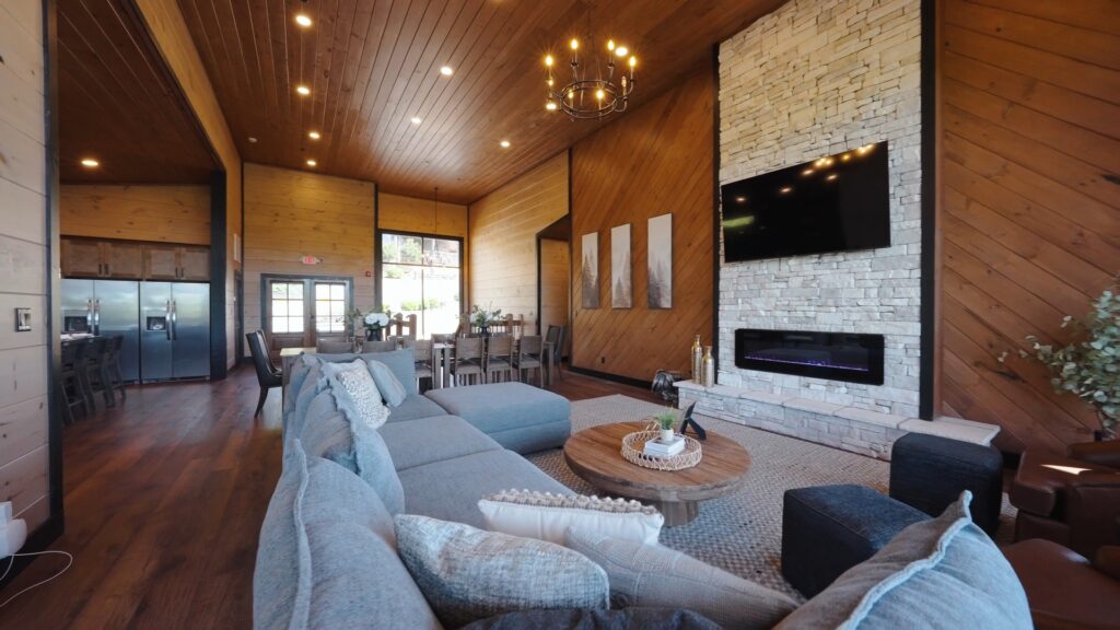 Side perspective of a cabin living room featuring comfortable couches, a FLOR type carpet, and a large flat-screen TV mounted over an electric chimney heater.