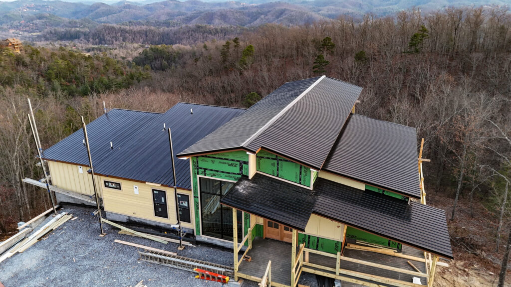 Aerial drone photograph of an active MCHB construction site in Sevierville, TN, showing a luxury cabin with finished roofing, structural heavy timber framing, and exterior glass windows installed.