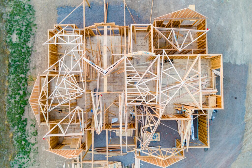 Zenith birds-eye-view aerial shot of a custom mountain home under construction, showcasing the raw lumber framing, floor joists, and architectural footprint.