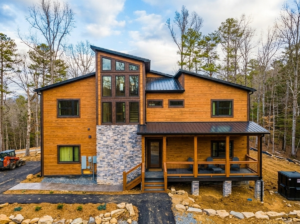 Side profile aerial drone view of a completed, fully furnished multi-level luxury mountain home by MCHB nestled in a lush green forest canopy in Sevierville, TN. Description: High-angle completed exterior portfolio photography documenting finished architecture, steep terrain site completion, multi-level exterior living decks, and landscape integration.