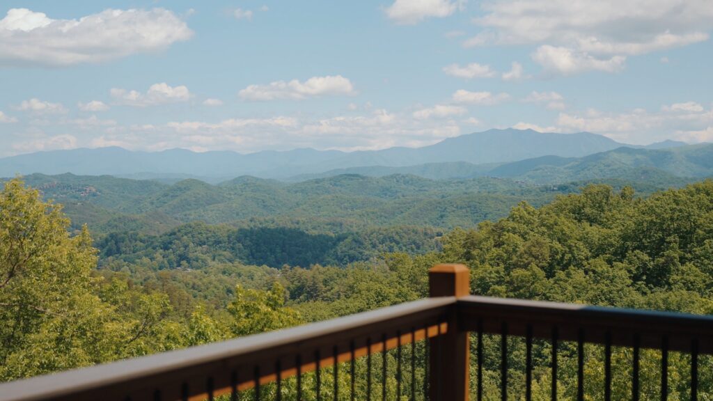 Panoramic view of the Smoky Mountain range and lush green forest under a blue sky from the balcony terrace of 3264 Laurel Cove Trail, showing a section of the deck railing.