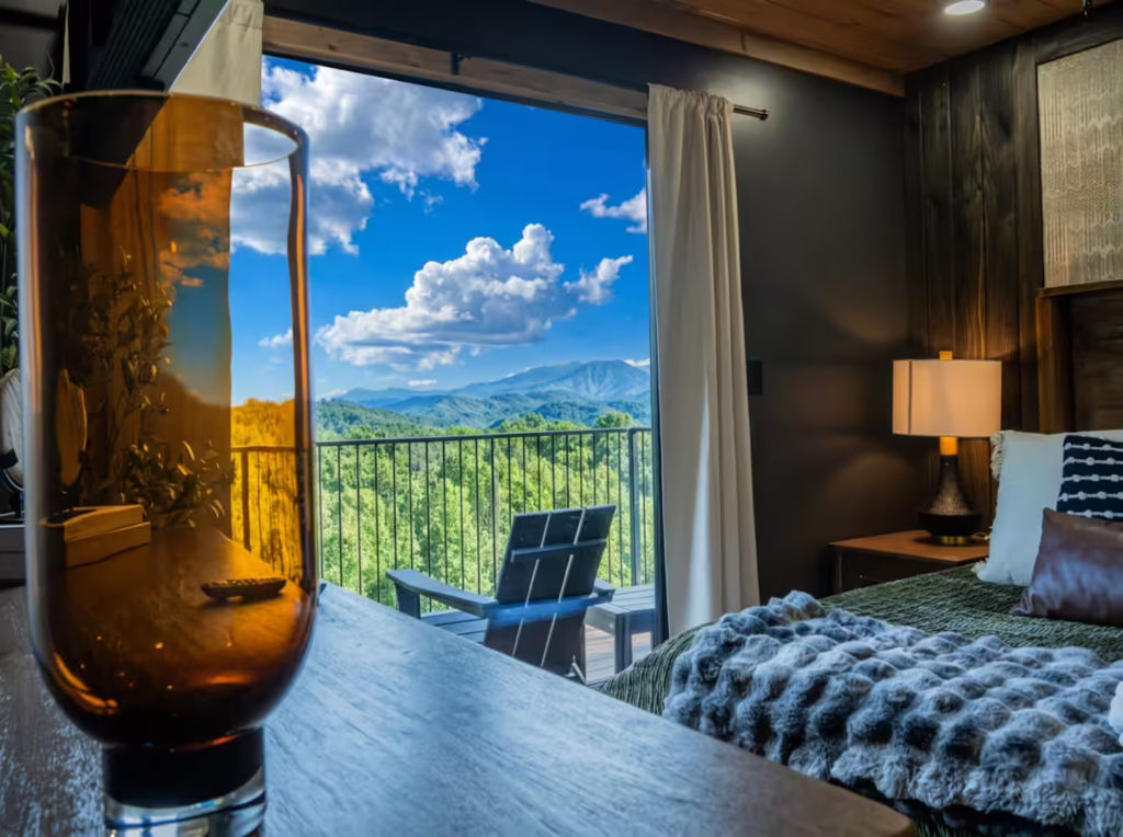Side angle view of a luxury master bedroom suite showing custom fine linens, premium modern furnishings, and a large picture window framing the blue ridges of the Smoky Mountains.