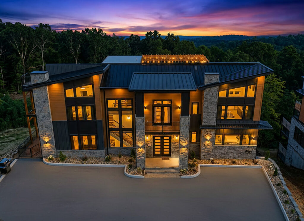 Majestic aerial drone twilight photograph of the completed 12-bedroom Reedmont Celeste Estate by MCHB, showcasing brilliant warm interior lighting through massive glass walls against a striking mountain sunset.