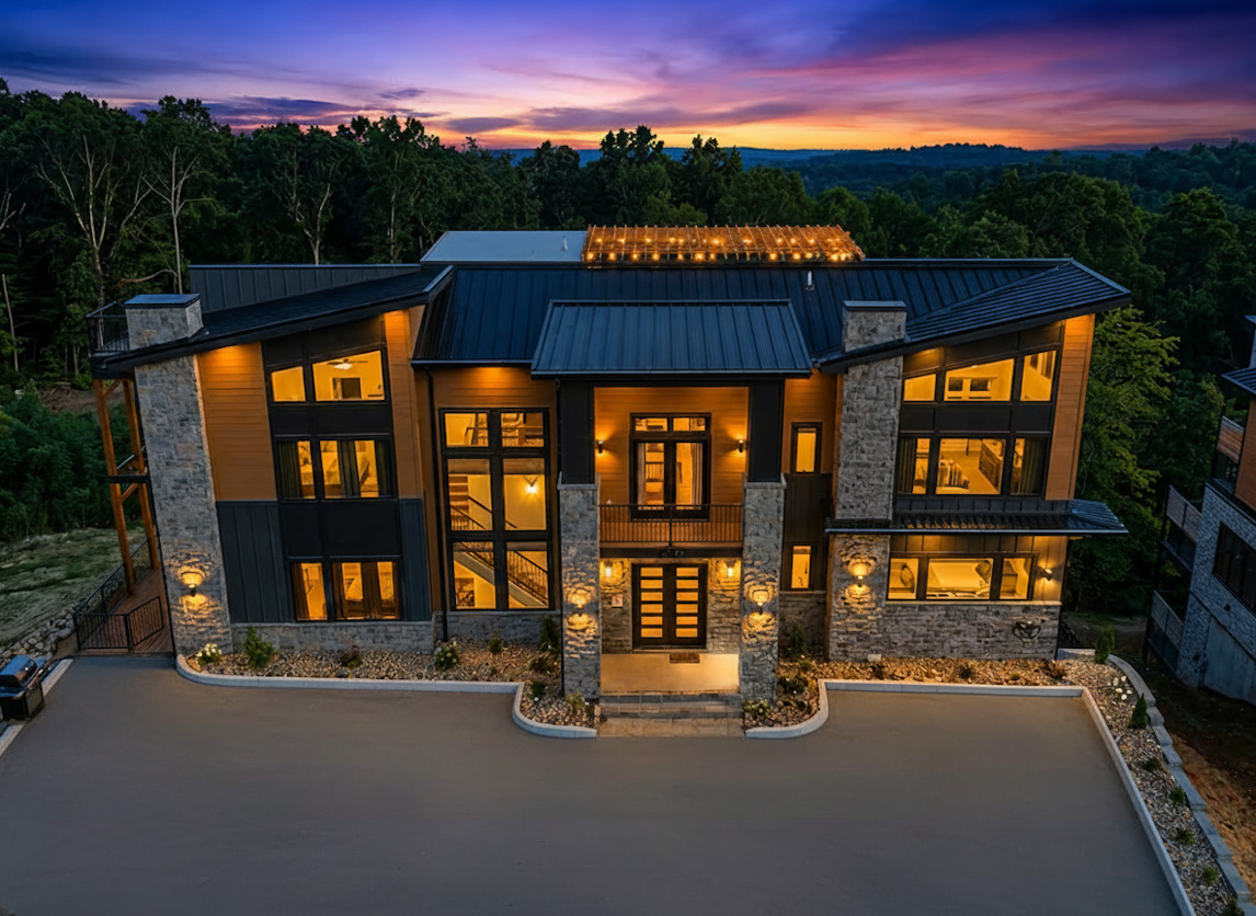 Majestic aerial drone twilight photograph of the completed 12-bedroom Reedmont Celeste Estate by MCHB, showcasing brilliant warm interior lighting through massive glass walls against a striking mountain sunset.