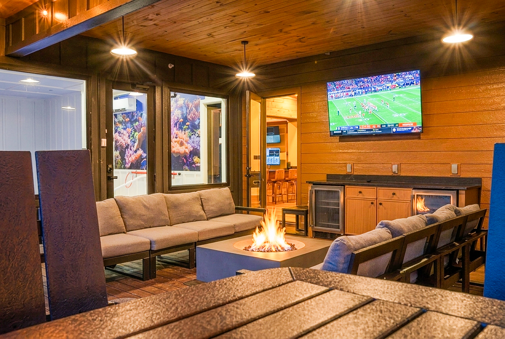 Outdoor deck terrace lounge with plush seating, outdoor stone fireplace, and wall-mounted TV, showing continuous view into an indoor entertainment station, game room, and heated indoor pool