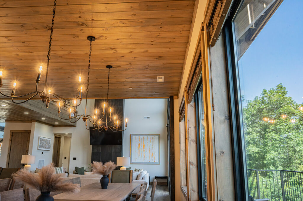 Low-angle tilt-up view of a luxury mountain home great room showing vaulted ceilings, custom designer lighting fixtures, open dining layout, and floor-to-ceiling mountain view windows.